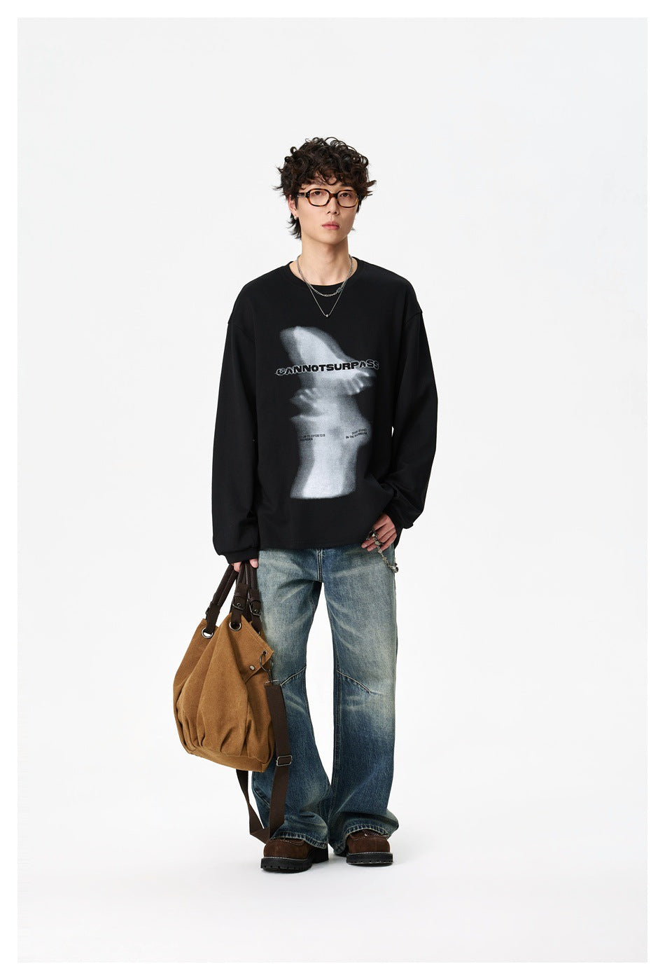 Full-length front view of a model wearing a black graphic sweatshirt, loose blue jeans, and brown shoes, holding a brown bag.