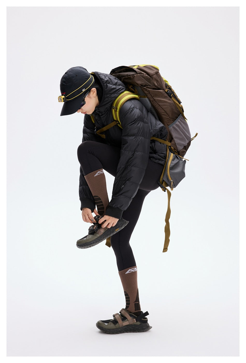 A model in a black puffer jacket and hiking backpack bends to adjust a hiking shoe.