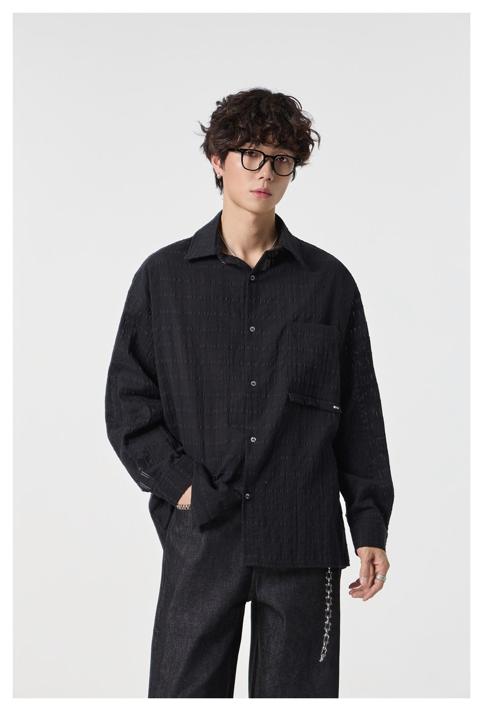Model wearing an oversized black textured button-up shirt with a chest pocket and dark jeans, shown from the front.