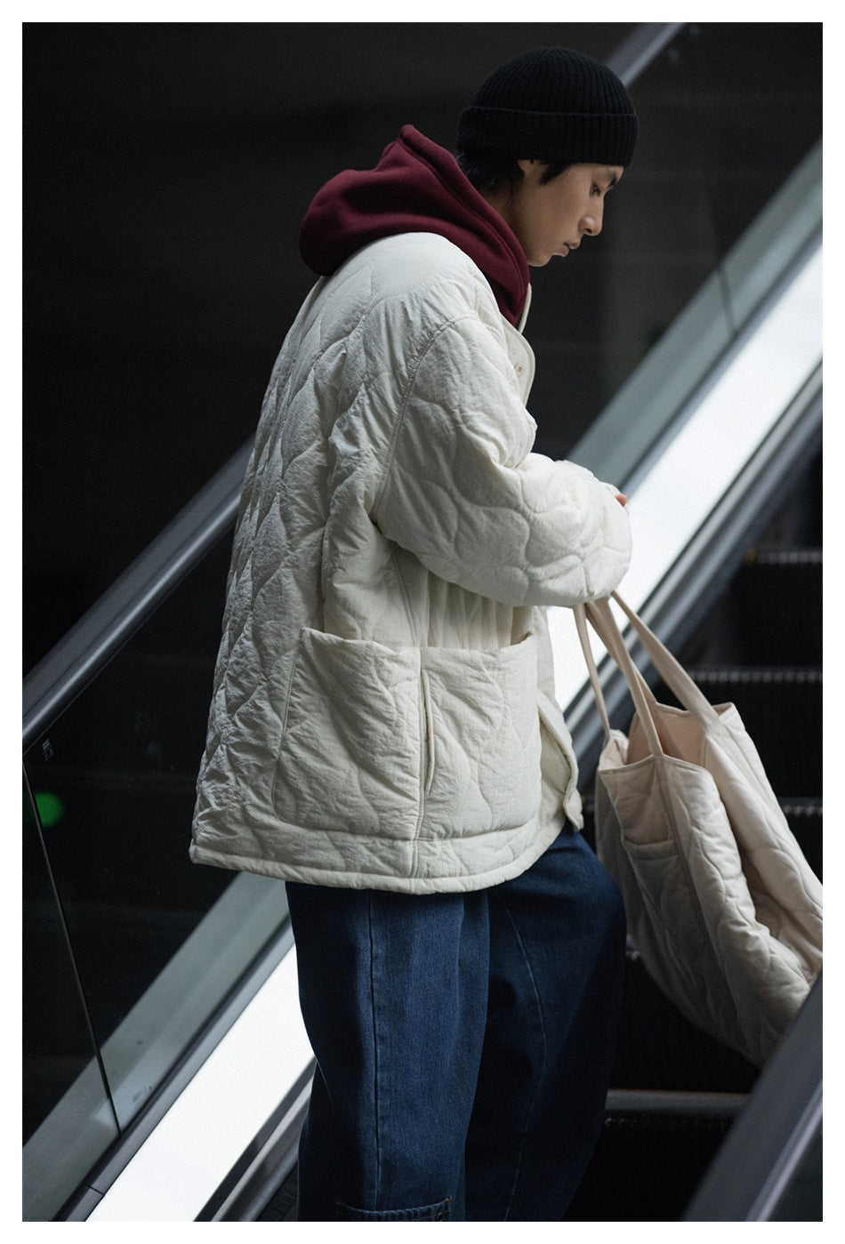 Side view of a person on an escalator wearing an off-white quilted jacket over a maroon hoodie, carrying a quilted tote