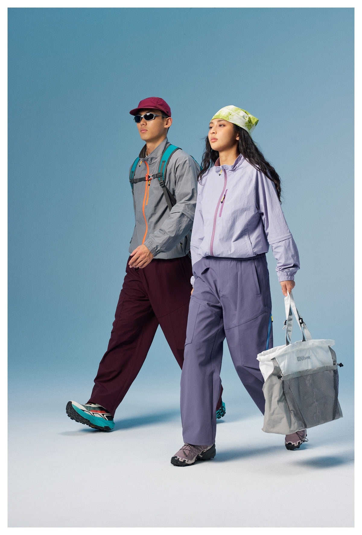 Two models walking in gray and lavender outdoor outfits, carrying backpacks and a tote bag.
