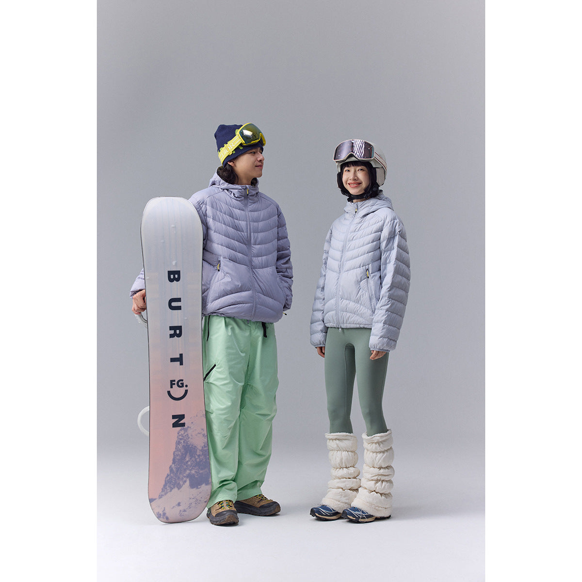 Two models wearing pale lavender hooded puffer jackets and ski pants, standing with a snowboard.