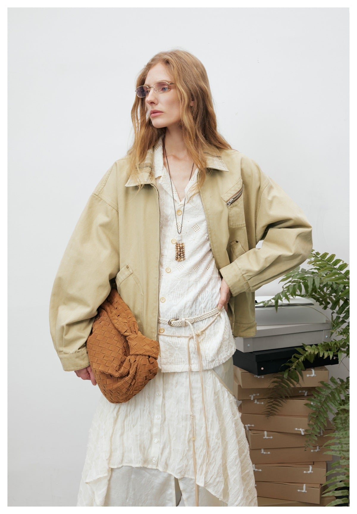 Close view of a model in a khaki zip jacket with plaid collar, holding a brown woven bag, hand on hip