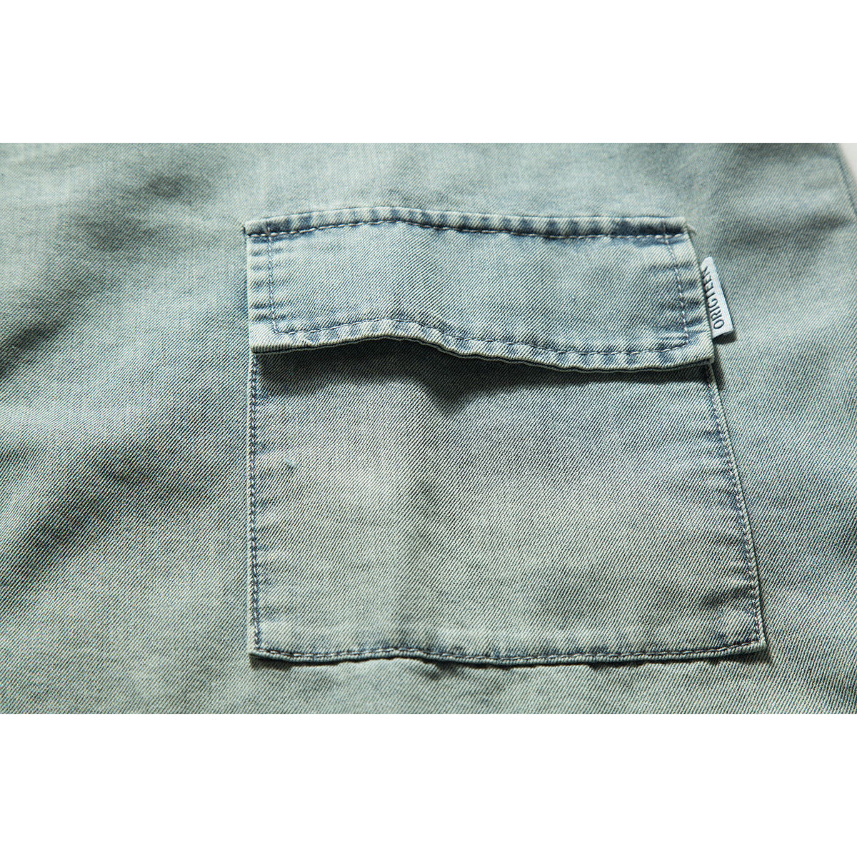 Close-up of a flap chest pocket on light blue denim, with a small sewn-in side label.