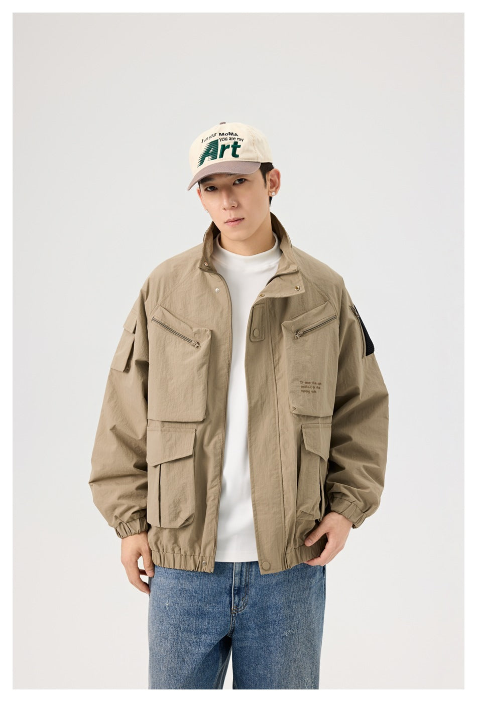 Model wearing an open khaki utility jacket with multiple pockets, a white shirt, blue jeans, and a cream cap.
