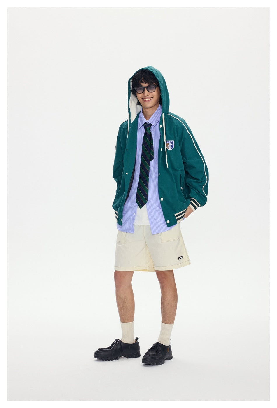 Smiling model wearing a dark green jacket with the hood up over a striped shirt, tie and cream shorts