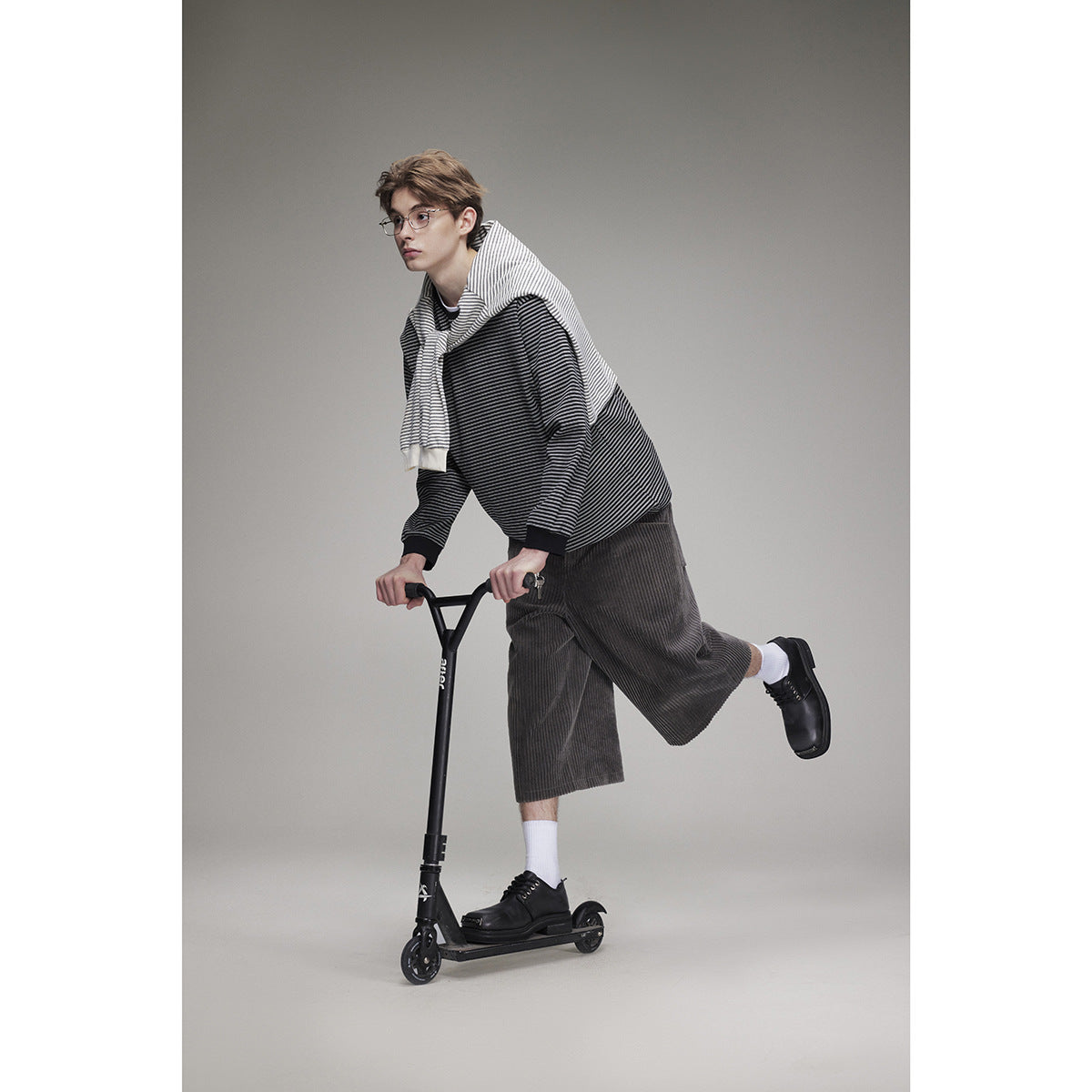 Model wearing a black-and-gray striped sweatshirt, matching draped top, and charcoal corduroy shorts on a scooter.