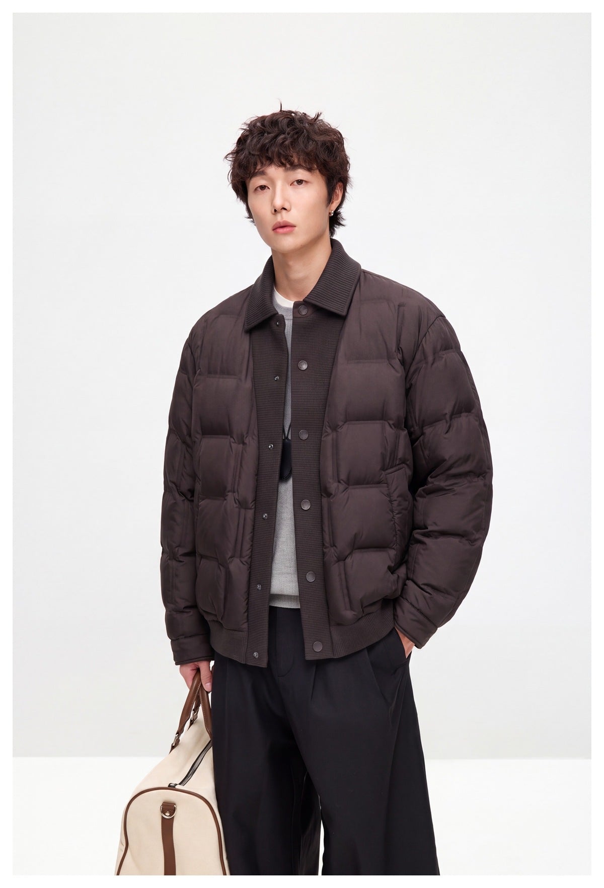 Model wearing brown quilted jacket over a gray top with black pants, holding a duffel bag, three-quarter view