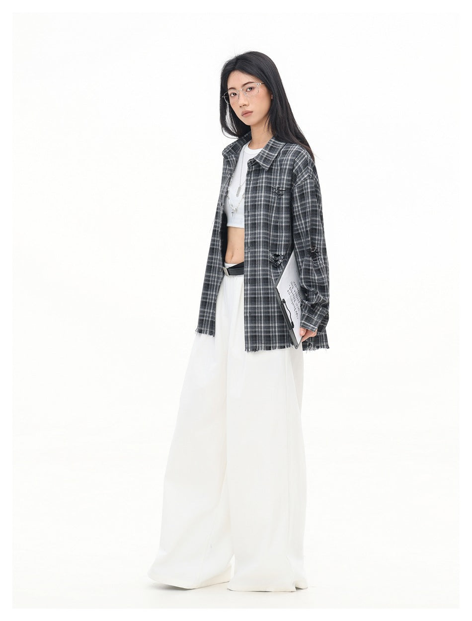 Model wearing open gray plaid distressed shirt over a white crop top with wide-leg white pants, holding a clipboard