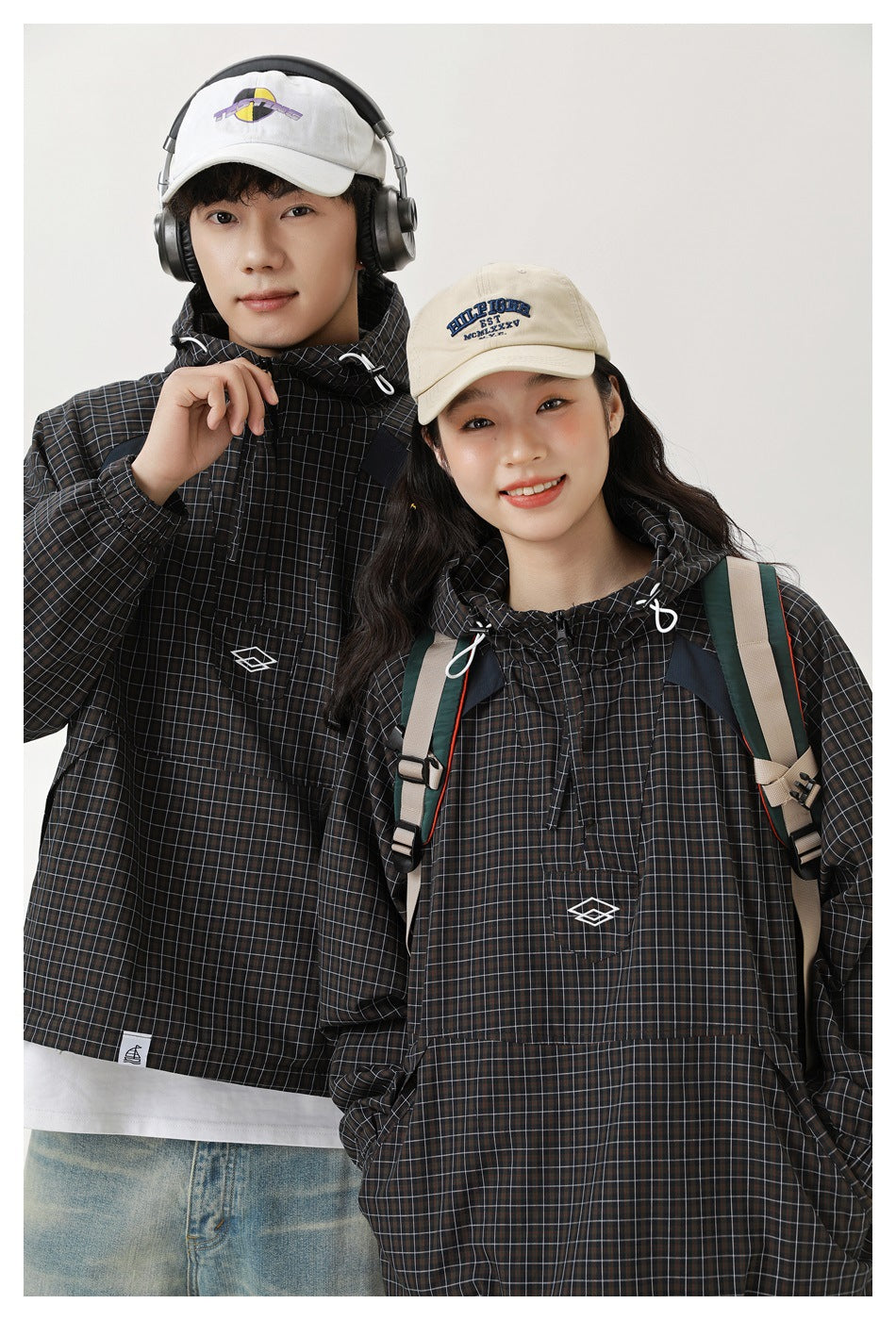 Two models wearing dark brown and black grid-plaid hooded pullover jackets with front pouch pockets.