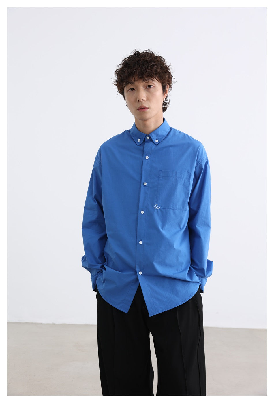 Model wearing a bright blue button-down shirt with a chest pocket and black trousers, shown from the front.