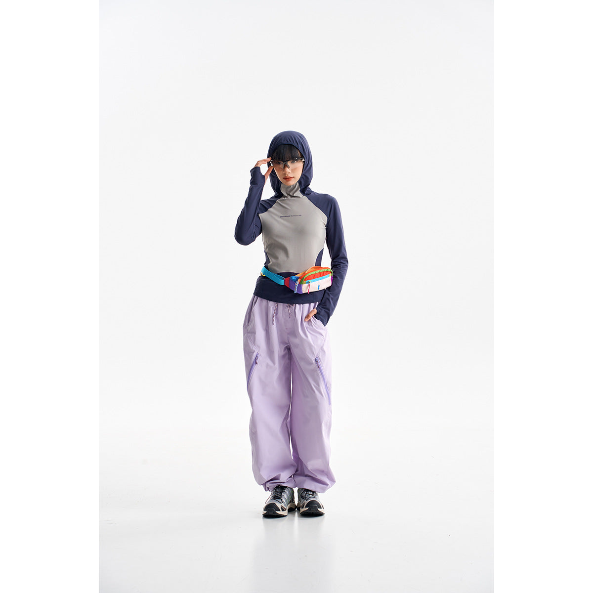 Model wearing a navy-and-gray hooded top, loose lavender pants, sneakers, and a colorful waist bag.