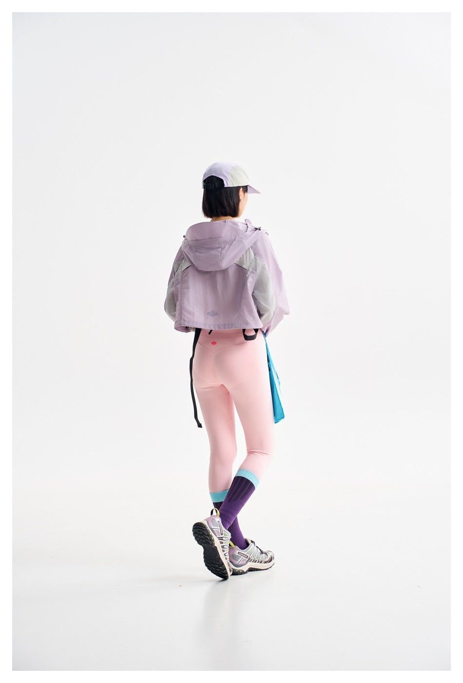 Rear view of a model wearing a cropped lilac hooded jacket with pale panels and pink leggings.