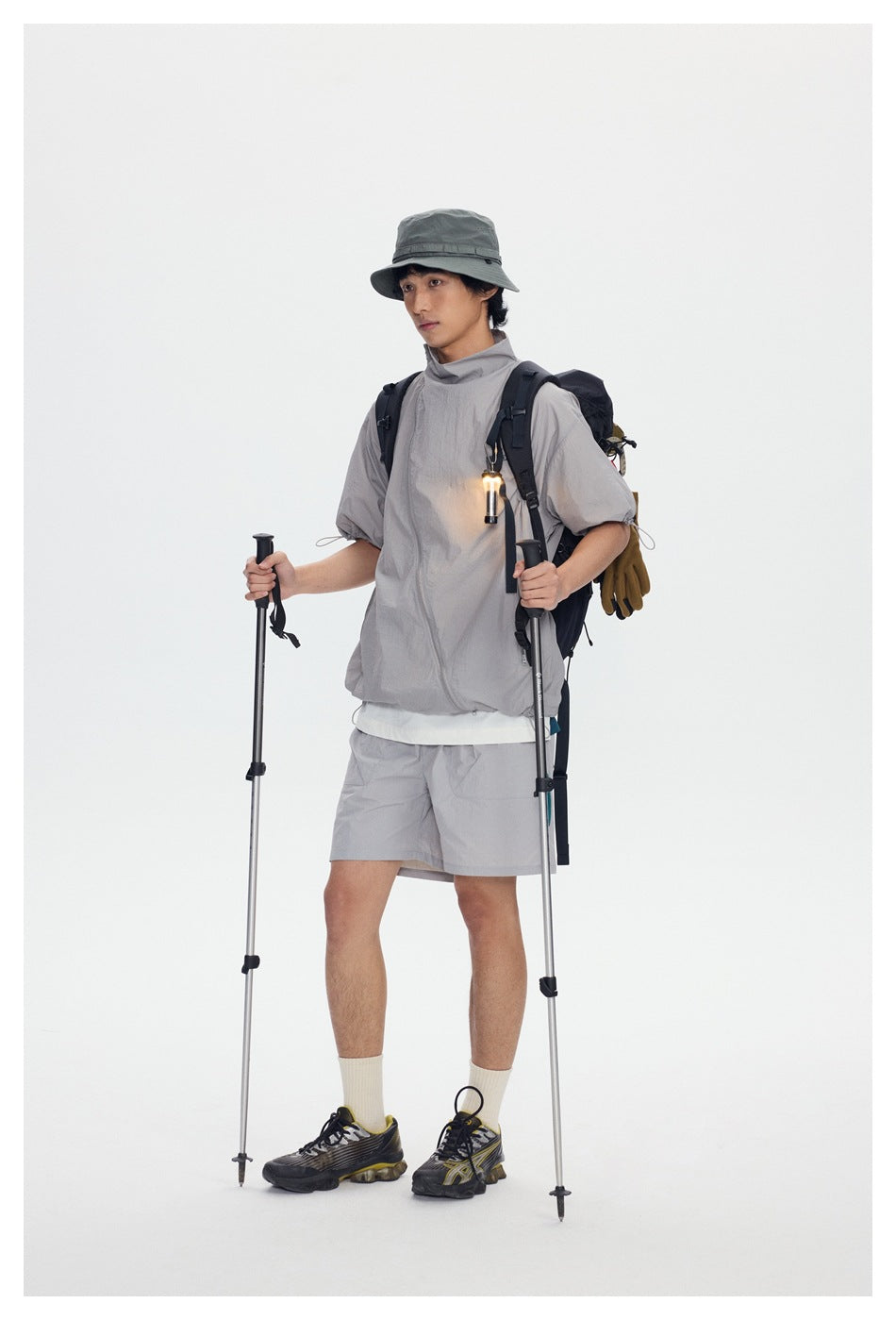 Full-length model in a light gray jacket and shorts with a bucket hat, backpack, and hiking poles.