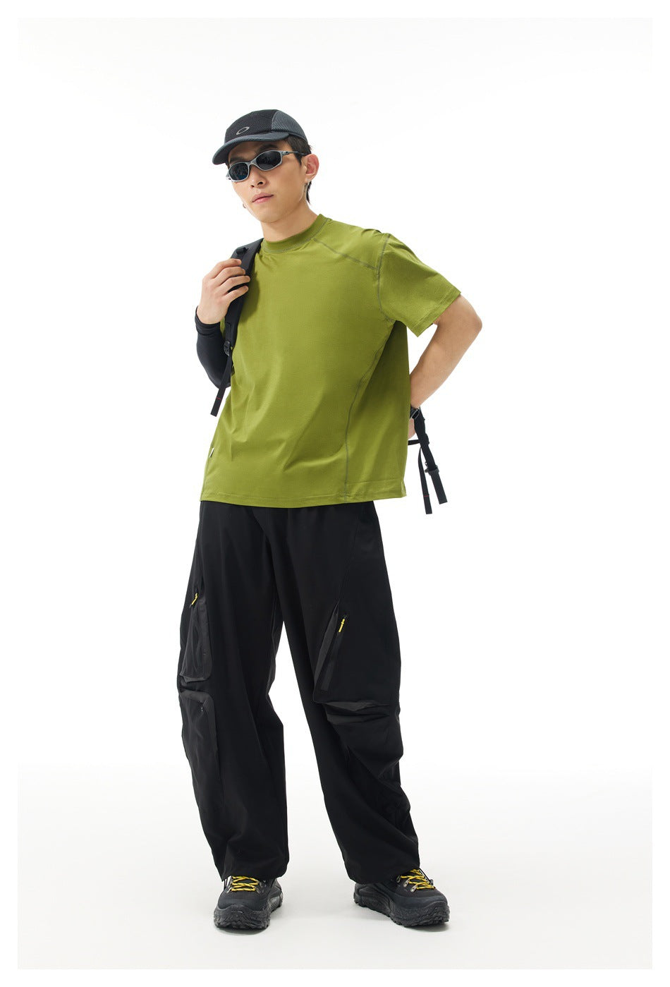 Full-length model wearing an olive-green short-sleeve shirt and black outdoor trousers with a backpack.