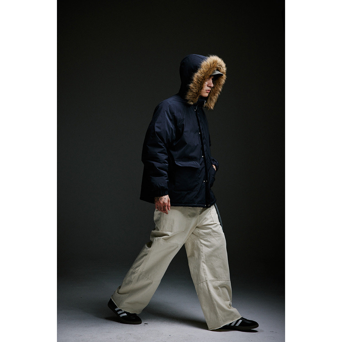 Full-length side view of a model in a navy fur-trimmed hooded parka and wide beige trousers.