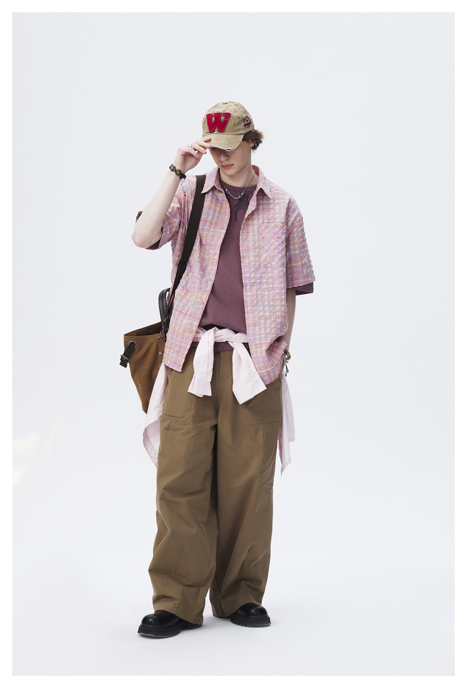 Full-length model adjusts his cap while wearing a pastel pink plaid short-sleeve shirt and olive trousers.