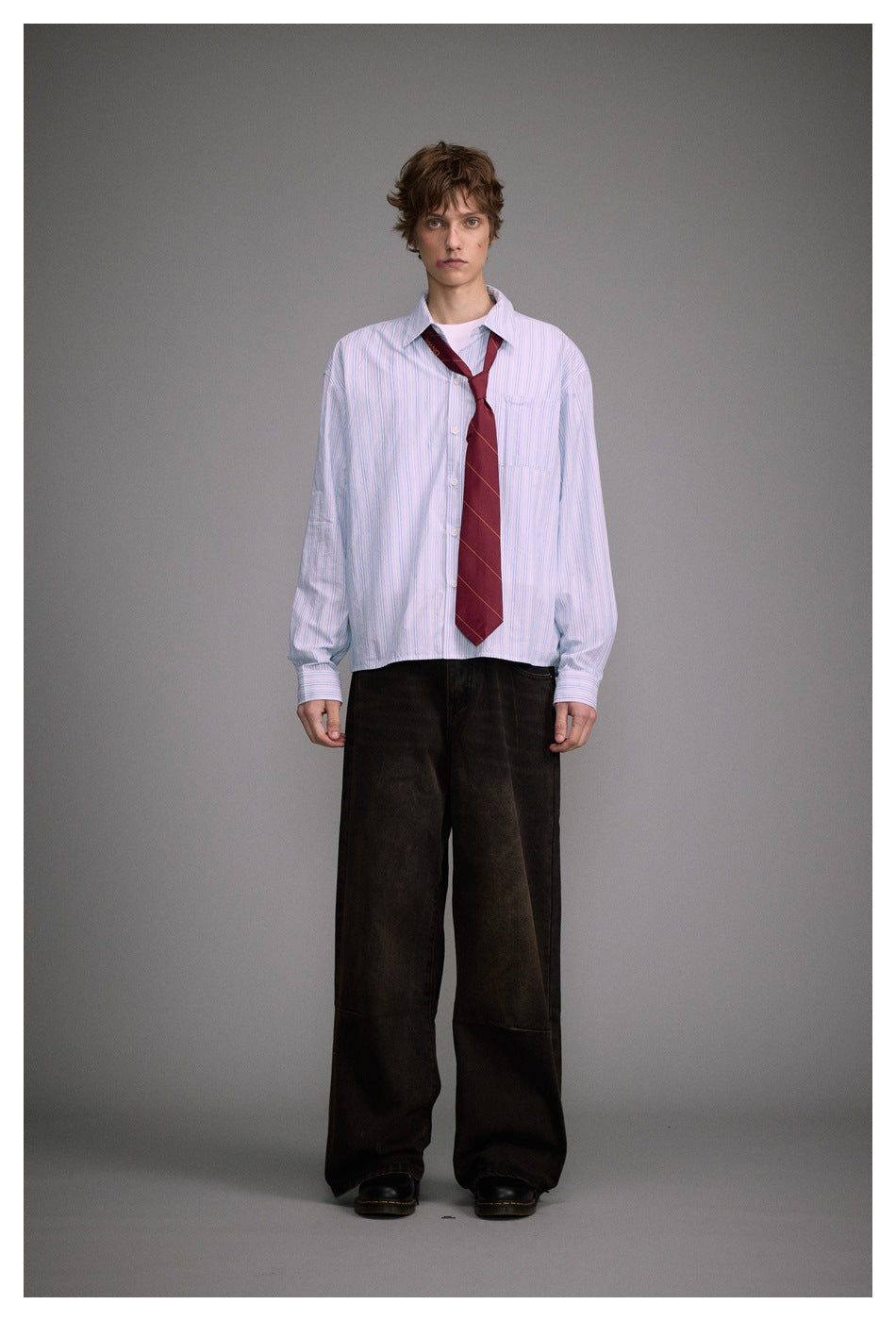 Model wearing the blue striped shirt open over a white tee with a red tie, dark wide-leg pants and black boots