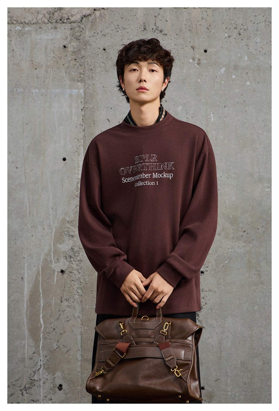 Model wearing a burgundy oversized crewneck sweatshirt with white chest text, holding a brown leather bag.