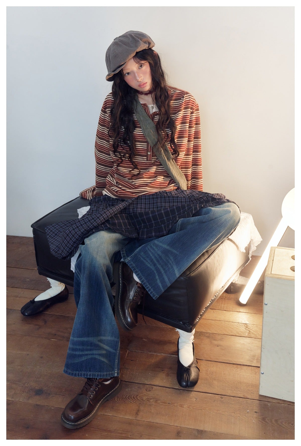 Model seated in a rust, brown and cream striped long-sleeve henley, wide-leg blue jeans and a navy plaid skirt.