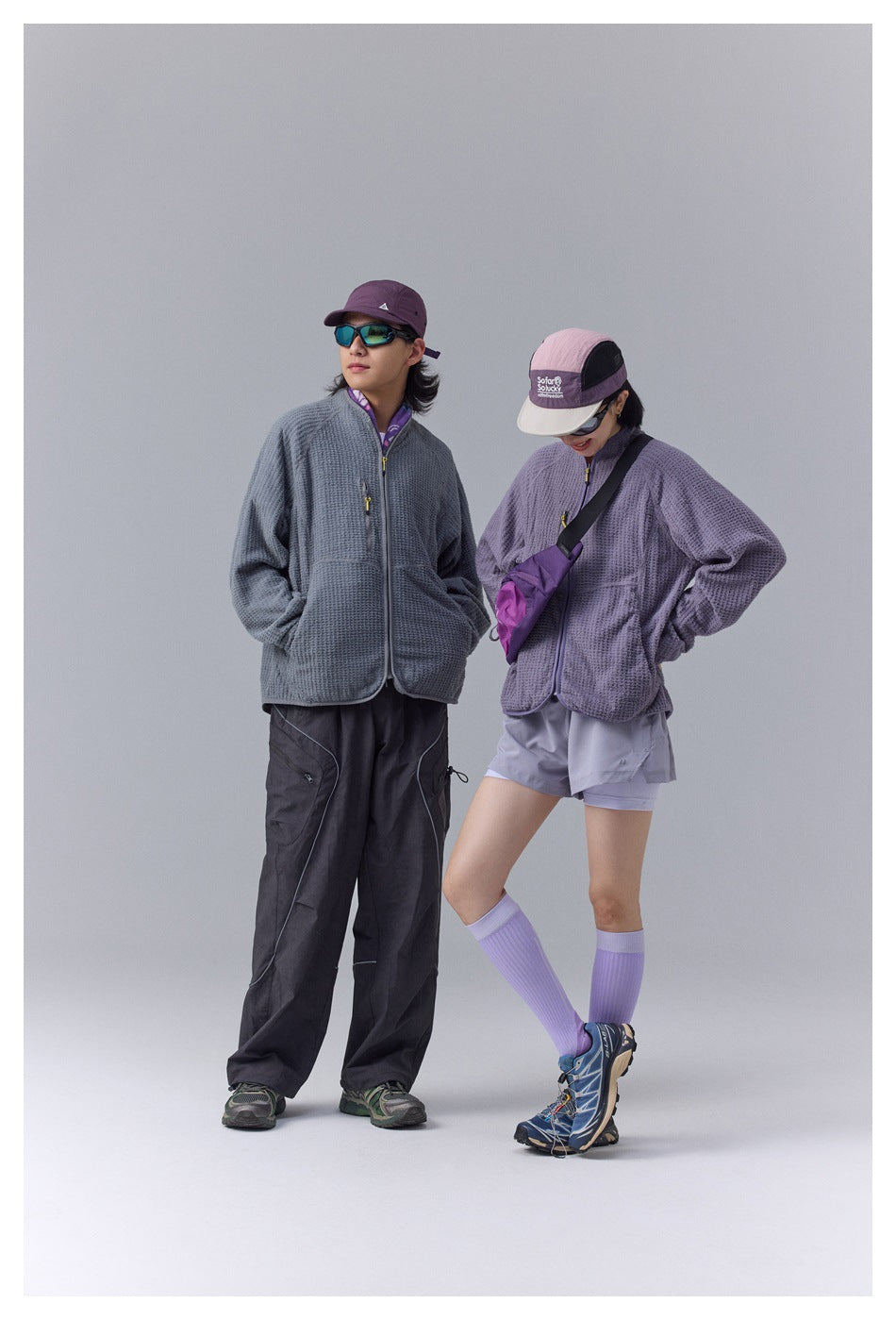 Two models wearing gray and purple textured zip jackets with caps, sunglasses, dark cargo pants and shorts, full-length pose
