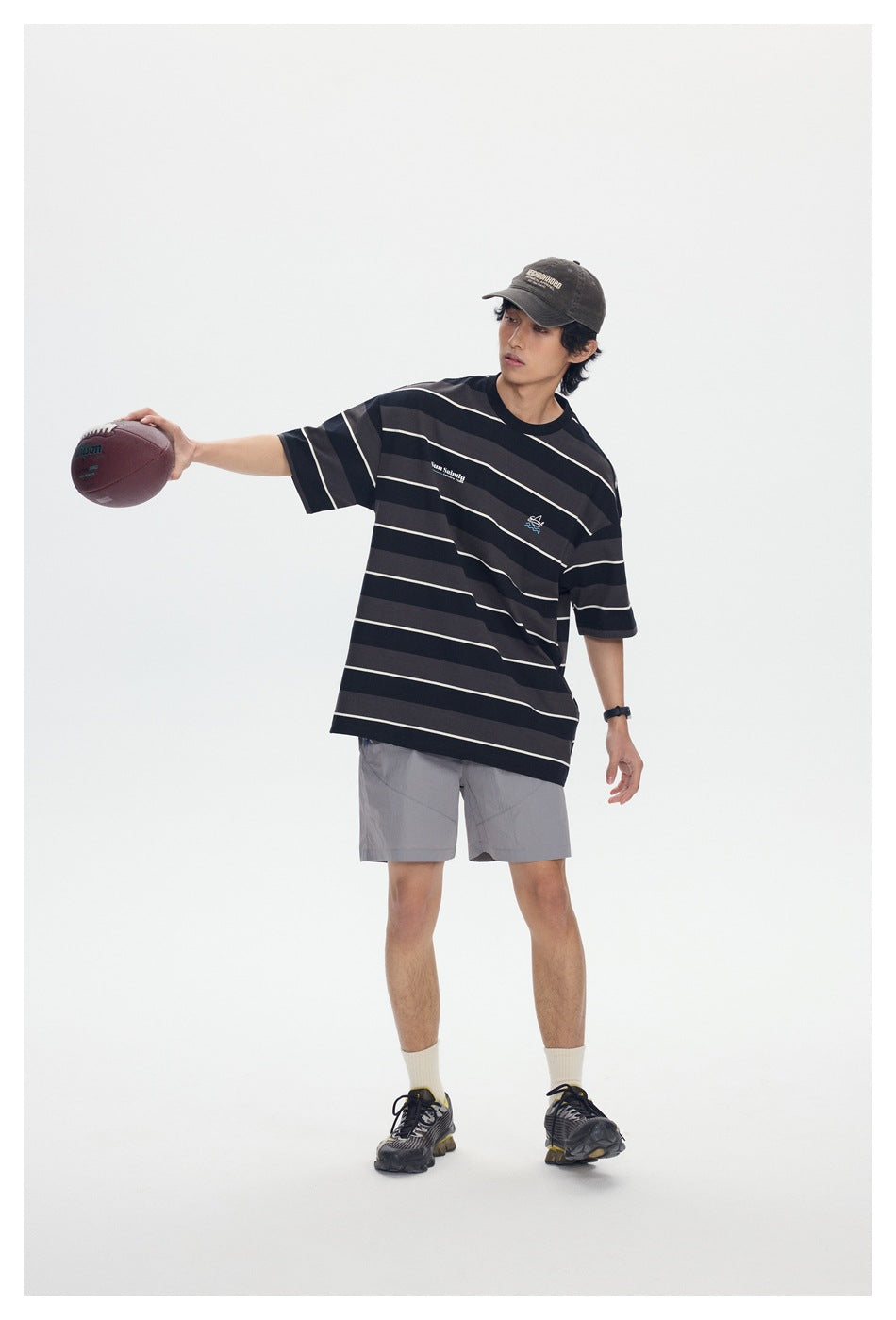 Model wearing a black and charcoal striped T-shirt and gray shorts, extending one arm with a football.