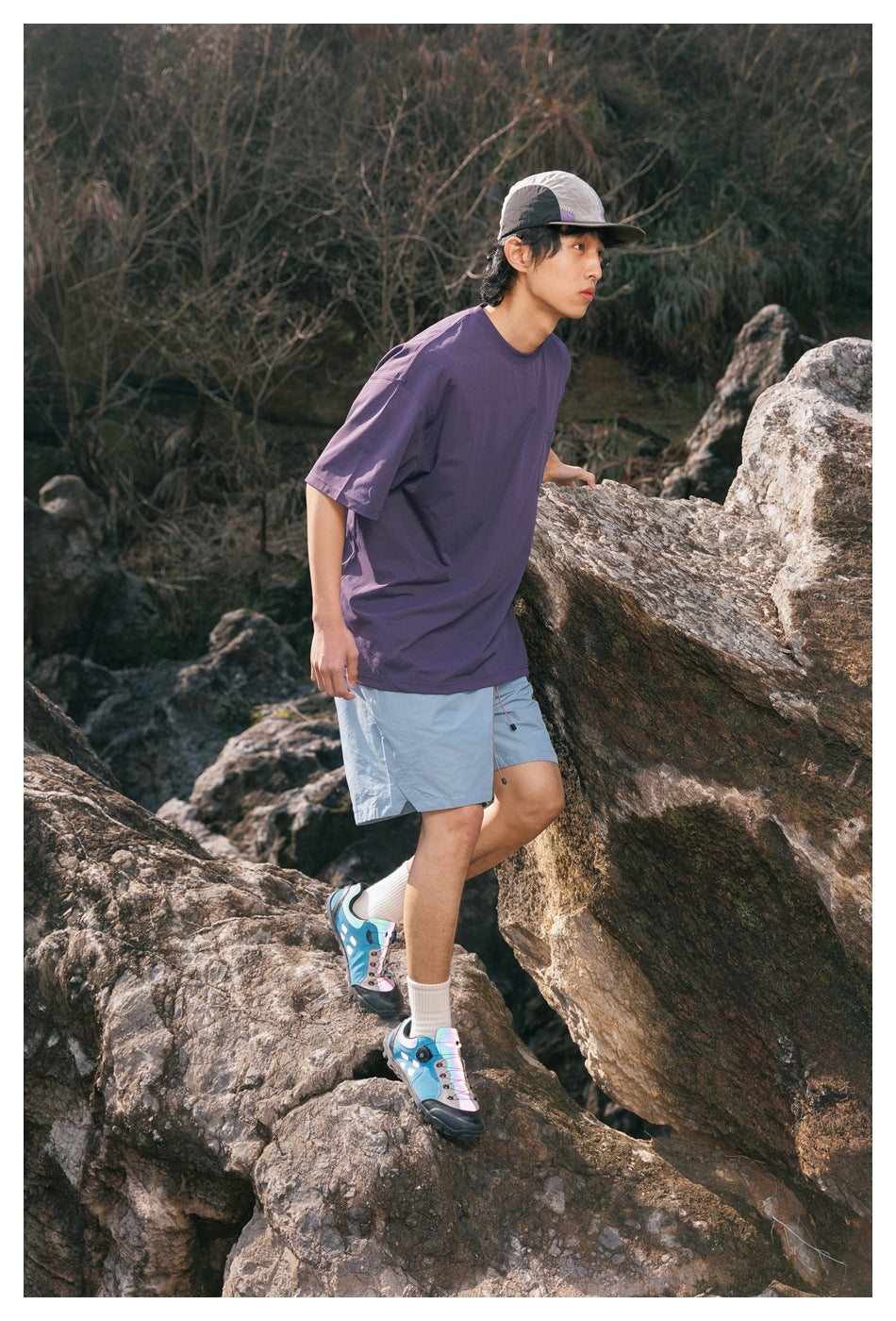 Model climbing rocks in a purple T-shirt, light blue shorts, and a two-tone cap.