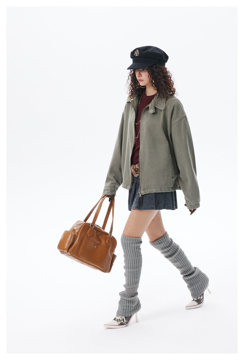 Model in an oversized olive-gray zip jacket, dark skirt, and gray leg warmers, carrying a brown handbag.