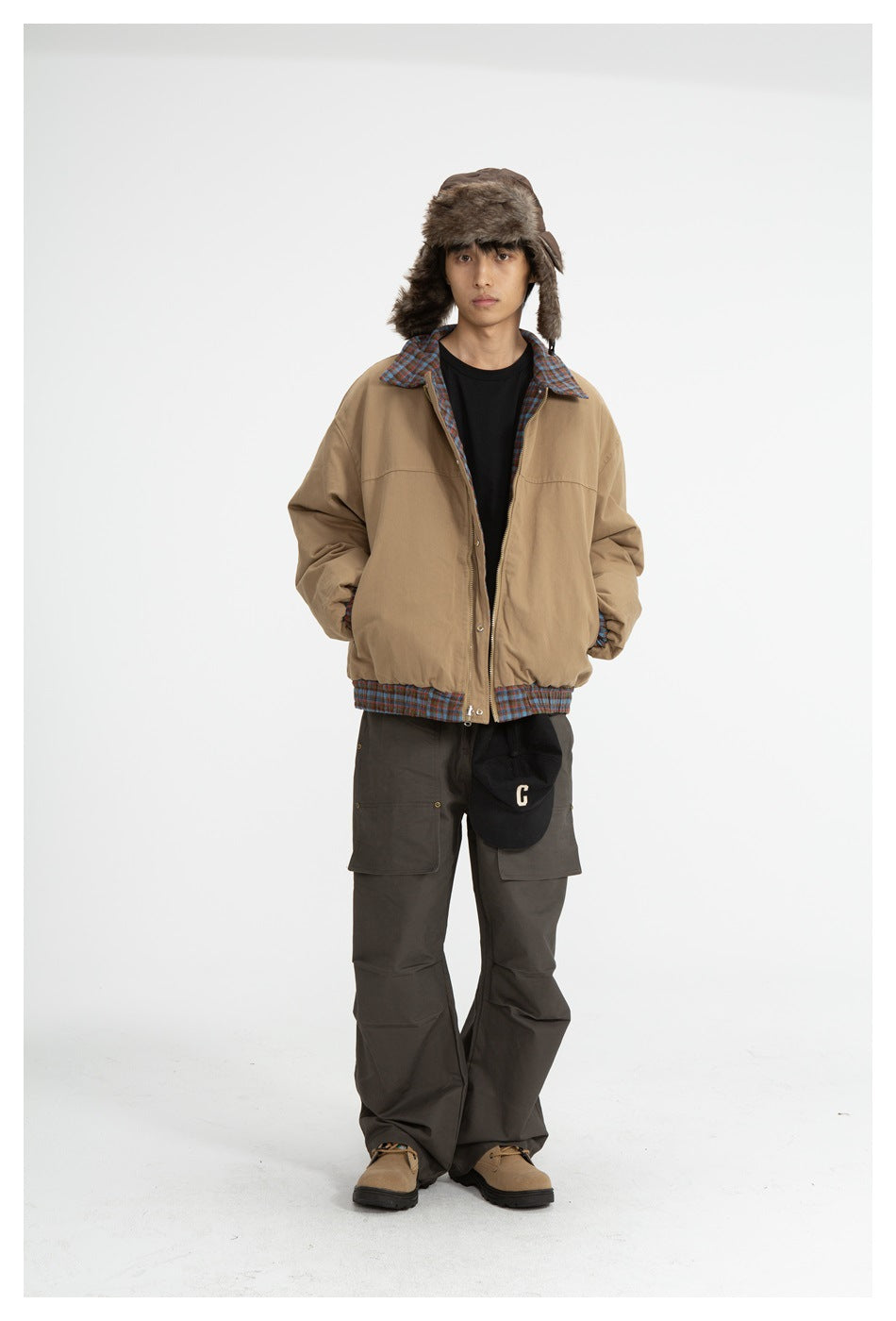 Model wearing open khaki jacket with plaid collar and hem over a black tee, brown cargo pants and a fur-style hat