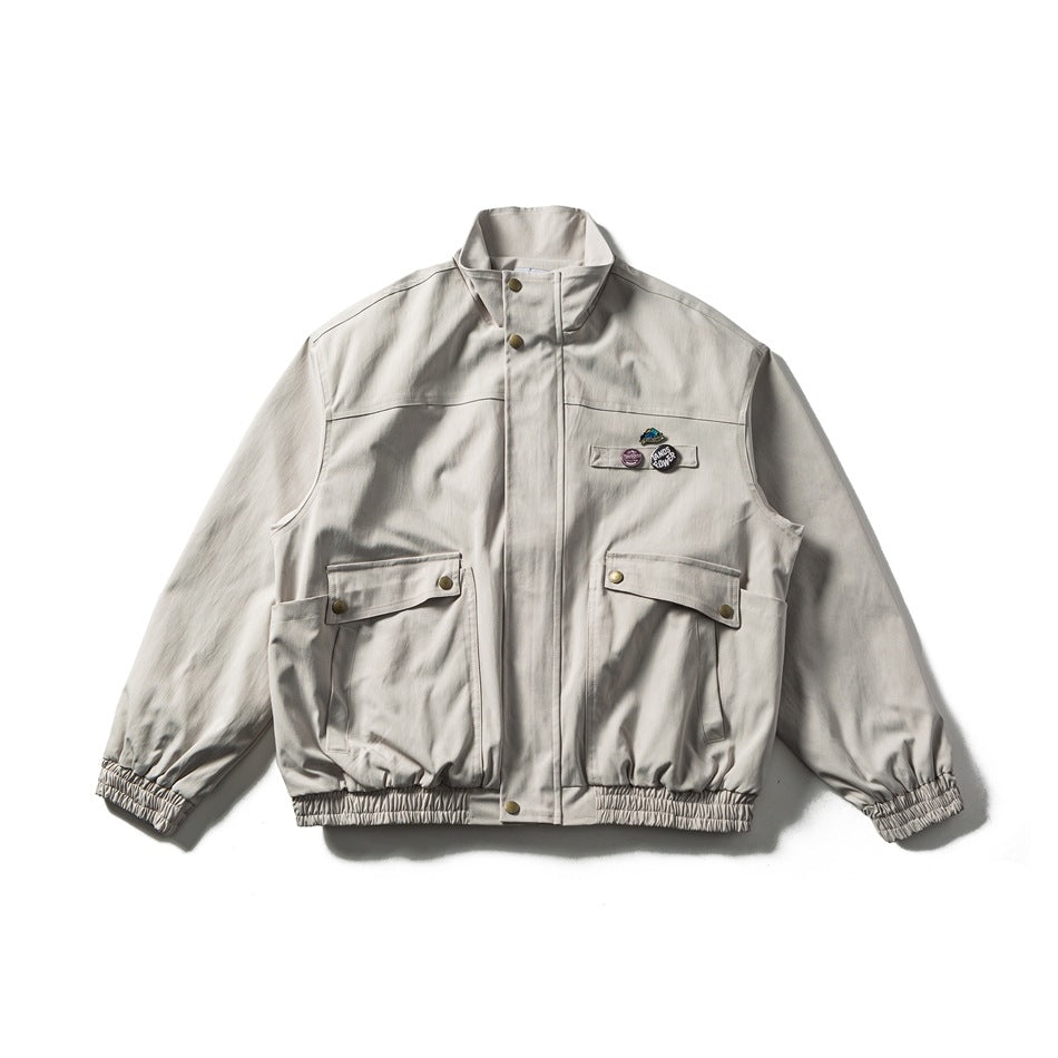 Light beige stand-collar jacket flat lay with two flap pockets, chest badges, and elastic cuffs and hem