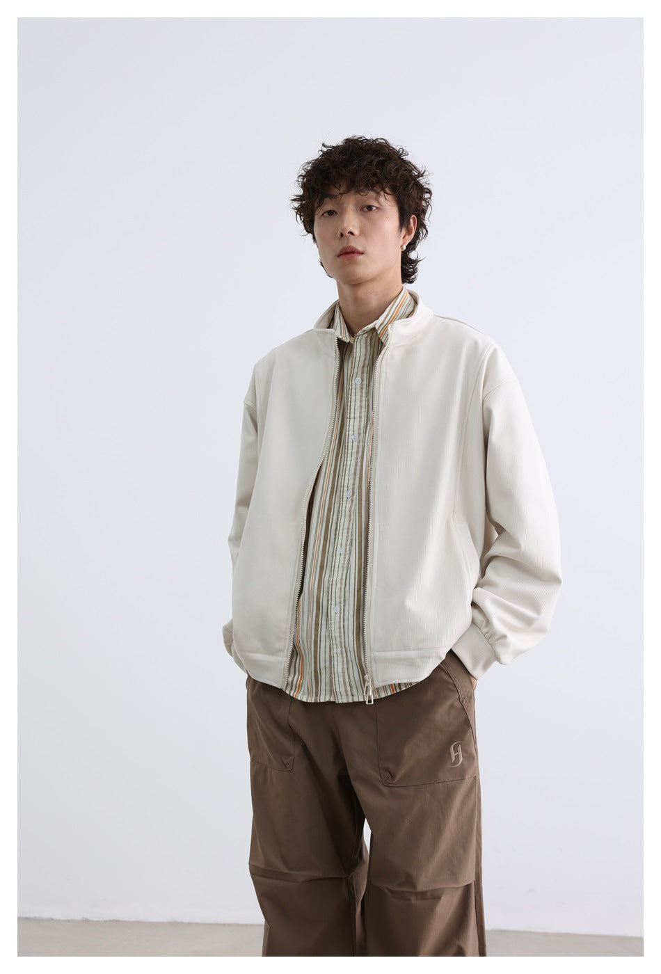 Model wearing an open off-white zip jacket over a striped shirt with taupe cargo-style trousers.