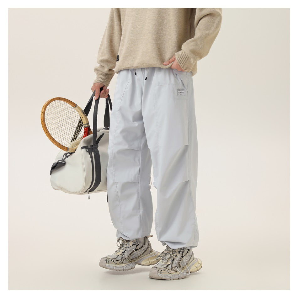 Front view of a model wearing very light gray loose pants with a side pocket, holding a tennis bag and racket.