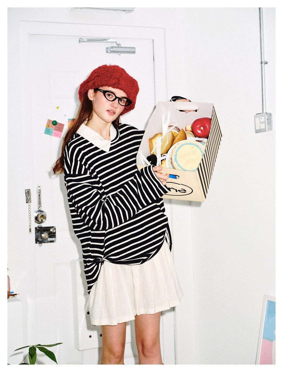 Model in black and white striped oversized top over a white collared shirt, red beret, holding a box of baked goods