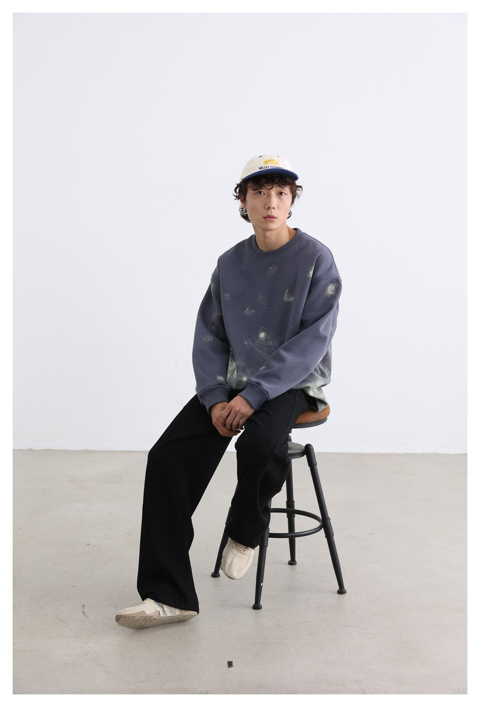 Model seated on a stool wearing a blue-gray printed sweatshirt with black pants and a cap