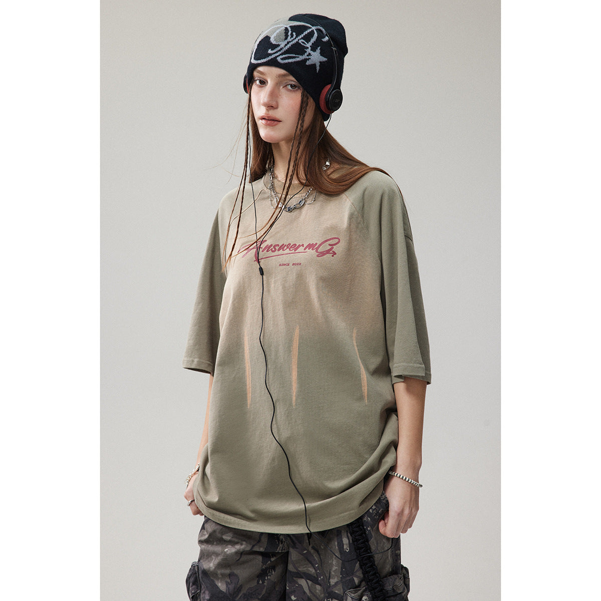 Model wearing an oversized olive-gray T-shirt with burgundy script and pale vertical streaks, paired with camouflage pants.