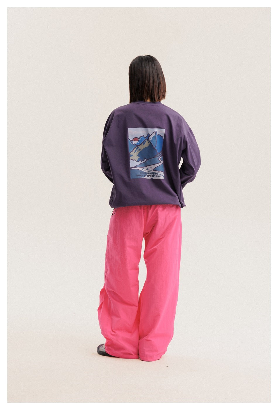 Rear view of a model wearing a plum long-sleeve T-shirt with a colorful mountain graphic and loose bright pink pants.