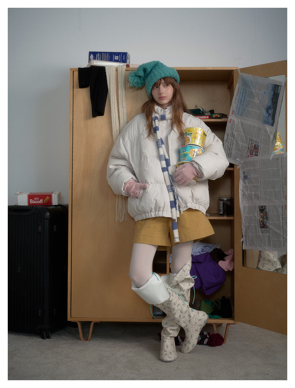 Model wearing white puffer jacket with striped scarf, teal pom hat, mustard shorts and boots beside a wooden wardrobe