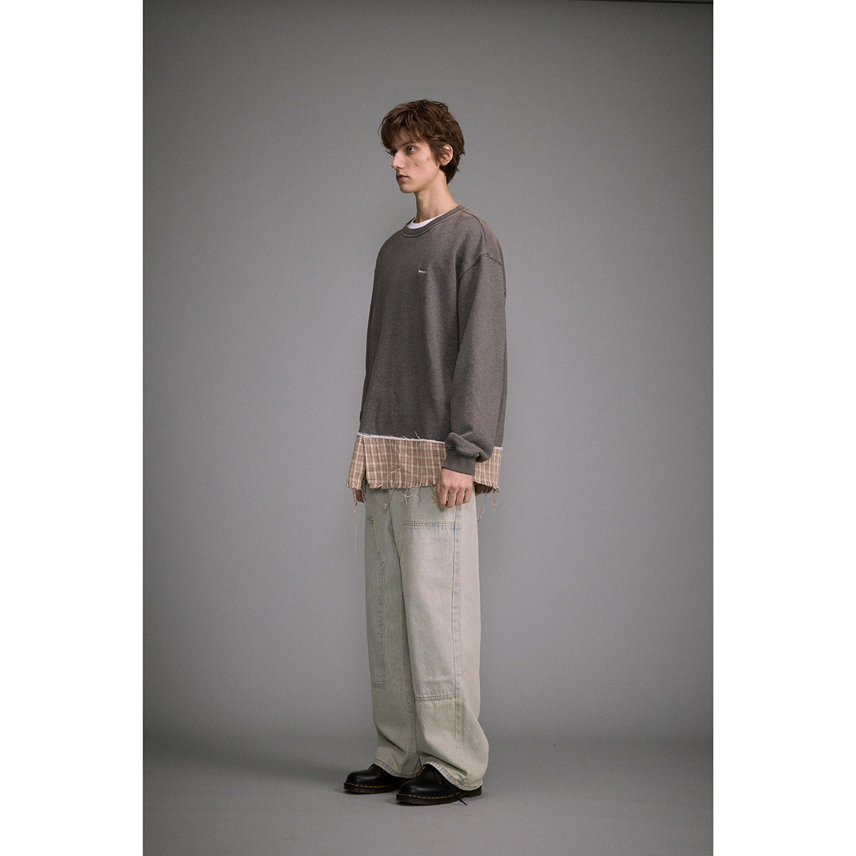 Man in a charcoal oversized sweatshirt with a frayed plaid hem, loose pale jeans, and black shoes, shown at an angle.