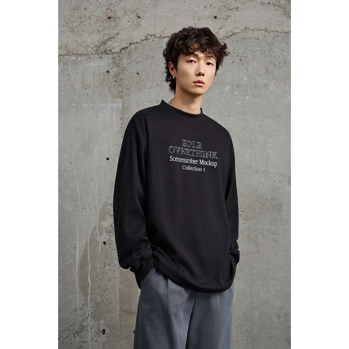 Front view of a model in a black oversized crewneck with white chest lettering and loose gray trousers.
