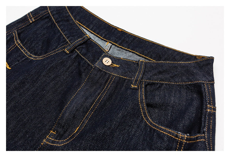 Close-up of dark indigo jeans showing the waistband, button, front pockets and golden contrast stitching.