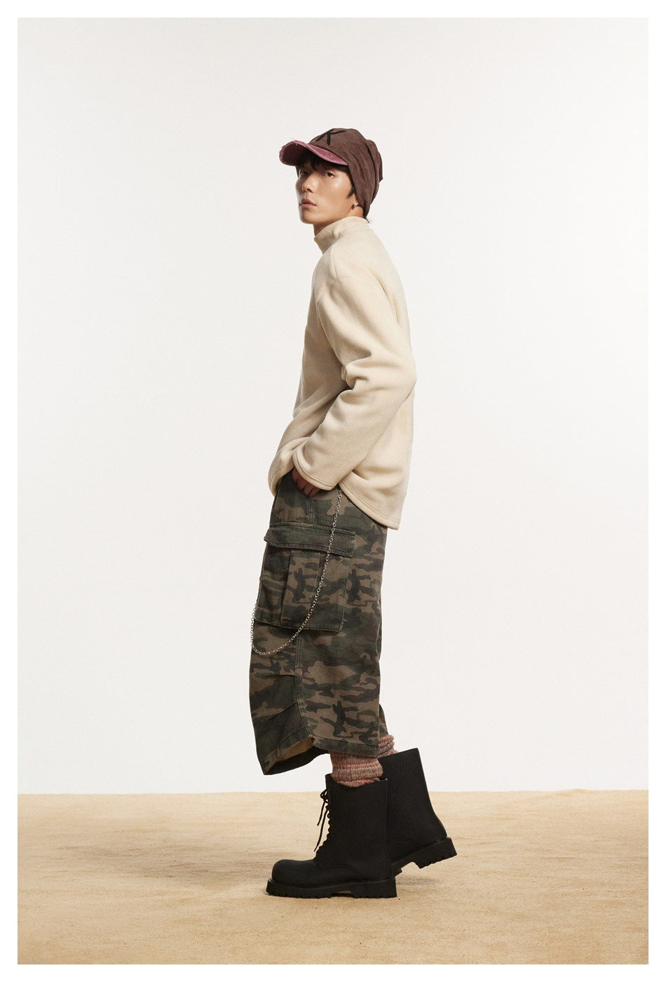 Side view of model in beige mock-neck pullover with camouflage cargo shorts, brown cap and black boots