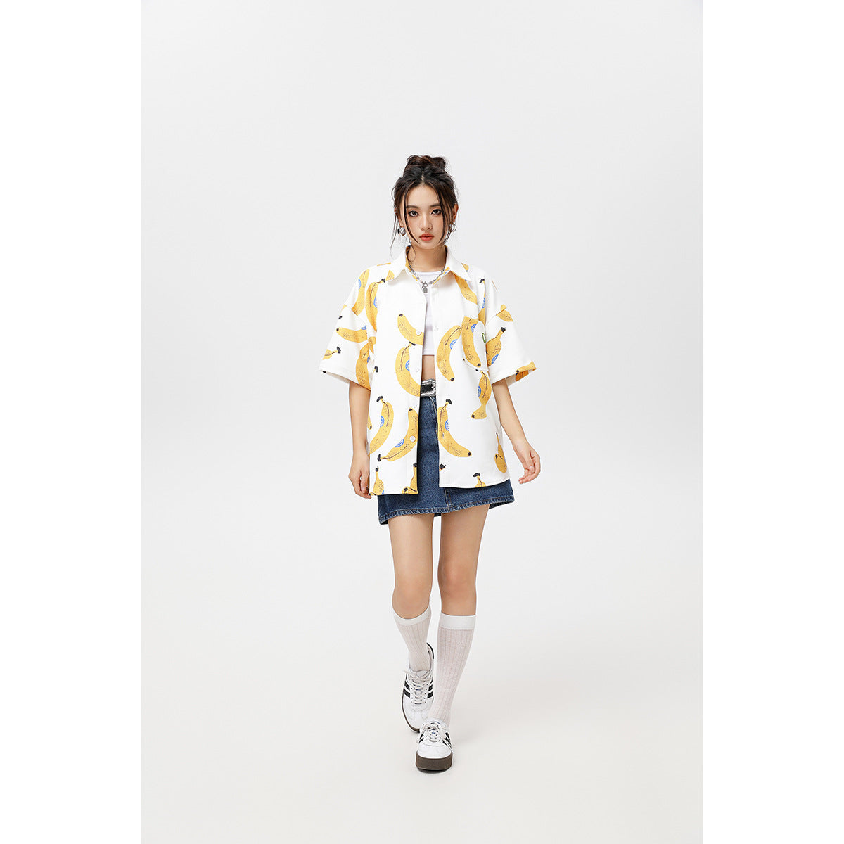 Model wearing an oversized white short-sleeve button-up shirt with a yellow banana print and a denim mini skirt.