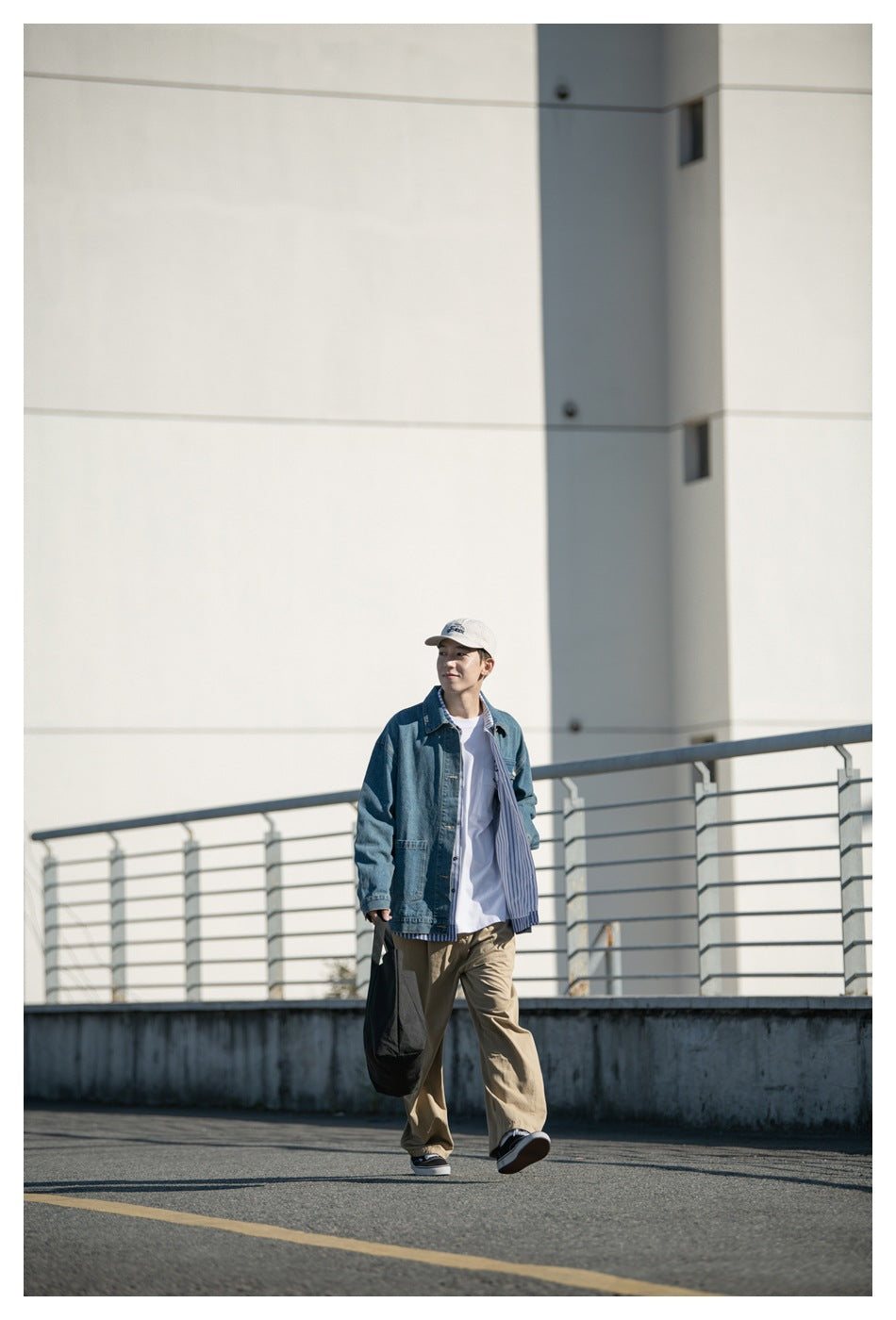 Model walking outdoors wearing an open blue denim jacket over a white tee, with khaki pants and a black tote bag