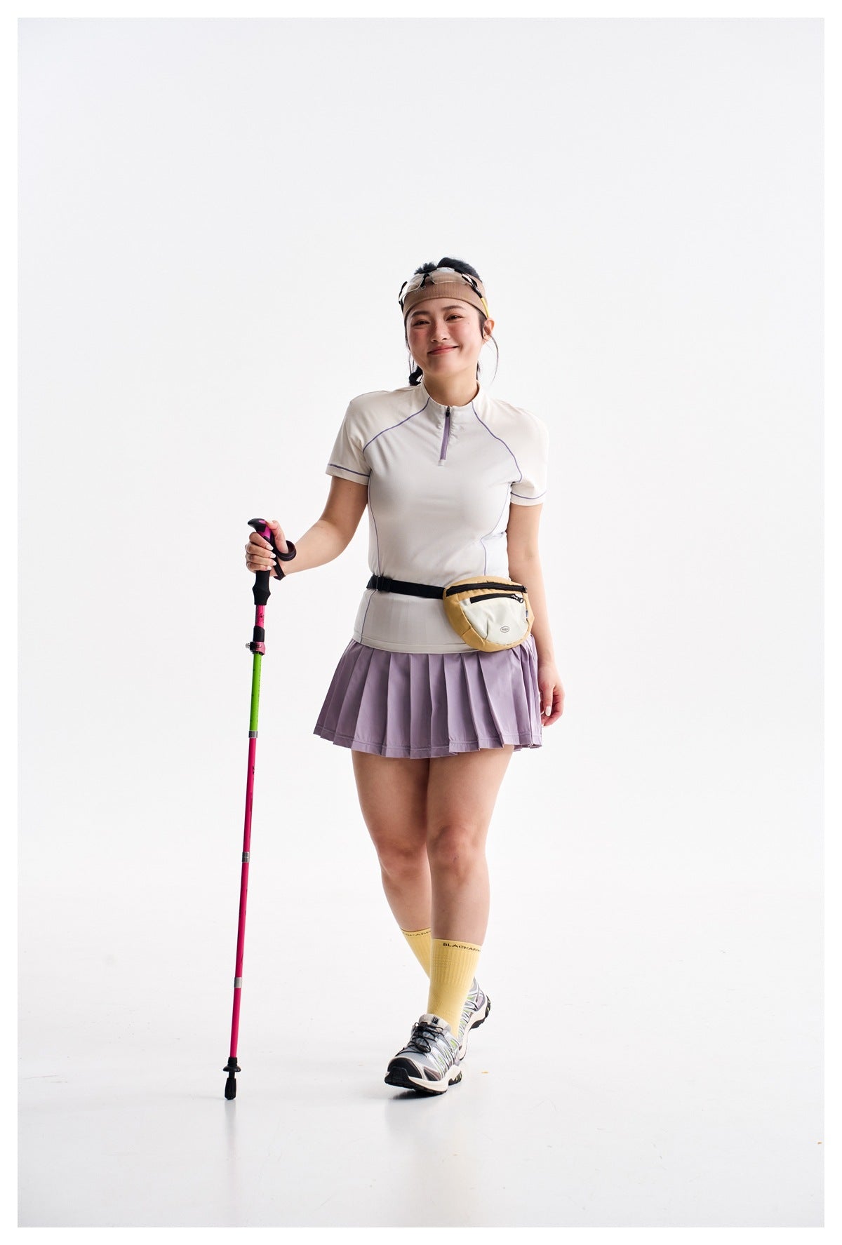 Model wearing a white short-sleeve zip top and pleated lilac skirt with a cream waist bag, holding a trekking pole.