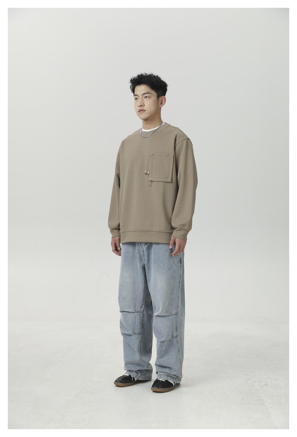Model in light brown crewneck sweatshirt with chest pocket and light wash jeans, angled three-quarter view