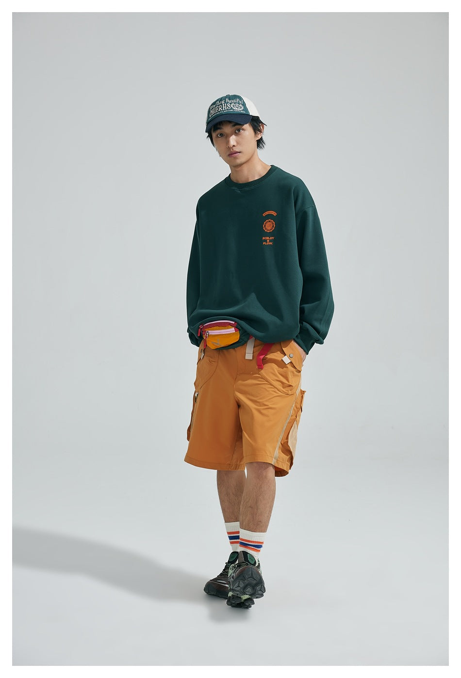 Full-length model wearing a dark green sweatshirt with orange chest print and ochre cargo shorts.