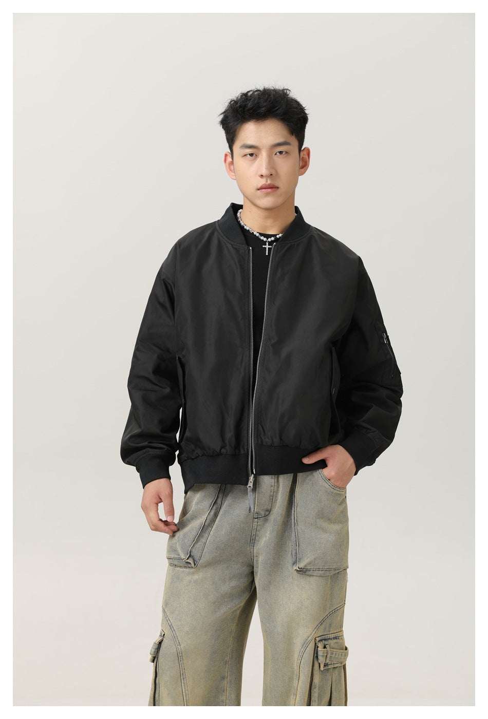 Man wearing a black bomber jacket with ribbed collar, cuffs and hem, unzipped over a black top and washed cargo jeans