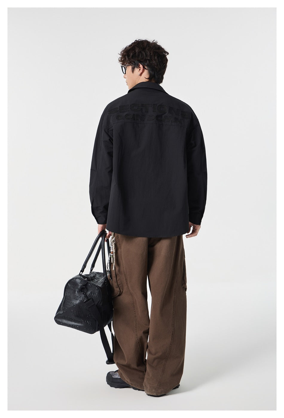 Back view of a model wearing a black button-up shirt with black lettering across the upper back and brown pants.