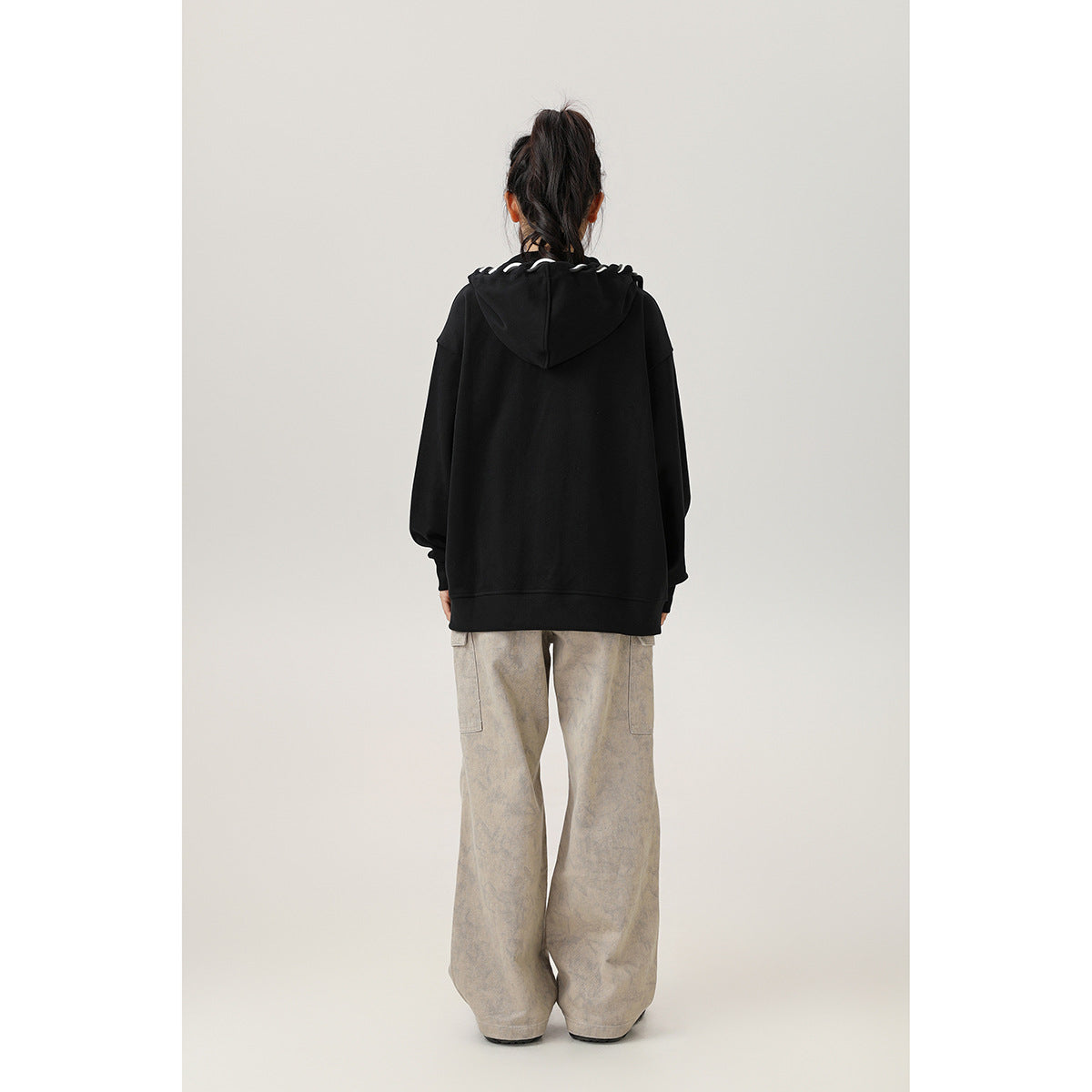 Back view of a model wearing an oversized black hooded sweatshirt with white trim at the hood and loose beige pants.