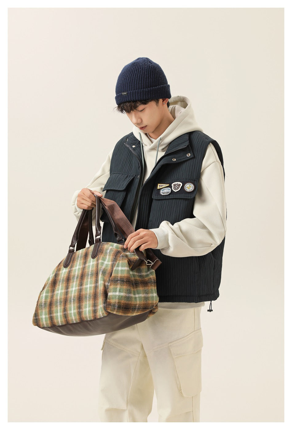 Model wearing a dark charcoal quilted utility vest with flap pockets and decorative patches over an ivory hoodie.